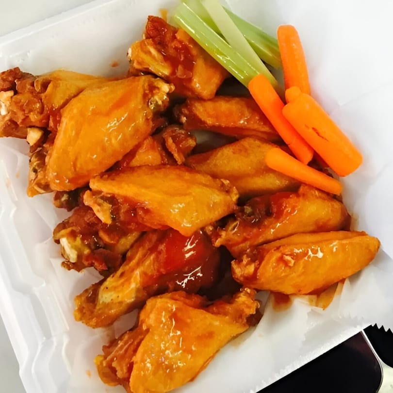 A white container filled with orange glazed chicken wings, celery sticks, and baby carrots.